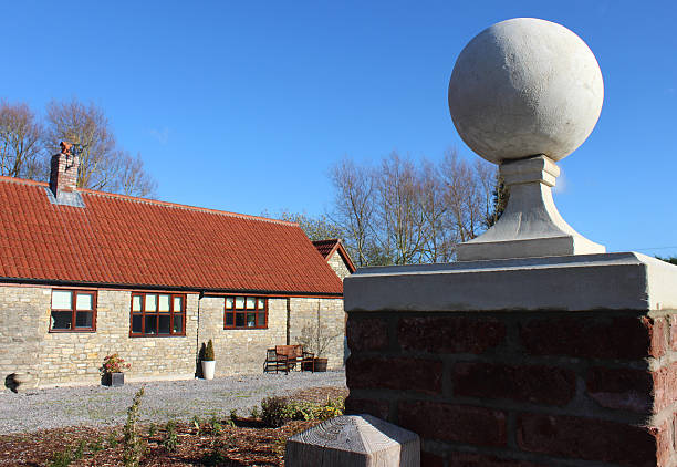 Photo showing a formal red brick gate post that has been topped with a pale moulded stone ball finial / pier cap, with a single-storey, barn conversion bungalow in the background (converted stables / outbuildings), complete with wooden porch and red roof tiles.  This stone ball is one of a pair, being positioned either side of a gate to create a particularly grand entrance.  The brick post has been photographed on a sunny afternoon, with a blue sky forming the natural background / backdrop.